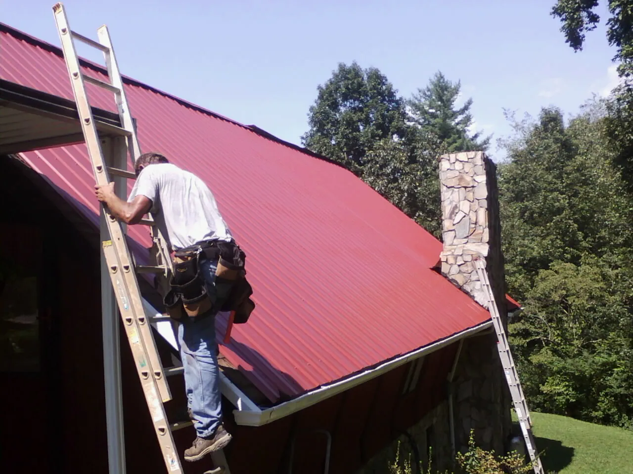 Roofer on a ladder beside a red metal roof and stone chimney — Soffit & Fascia Repair by Roofing Brothers in Munster